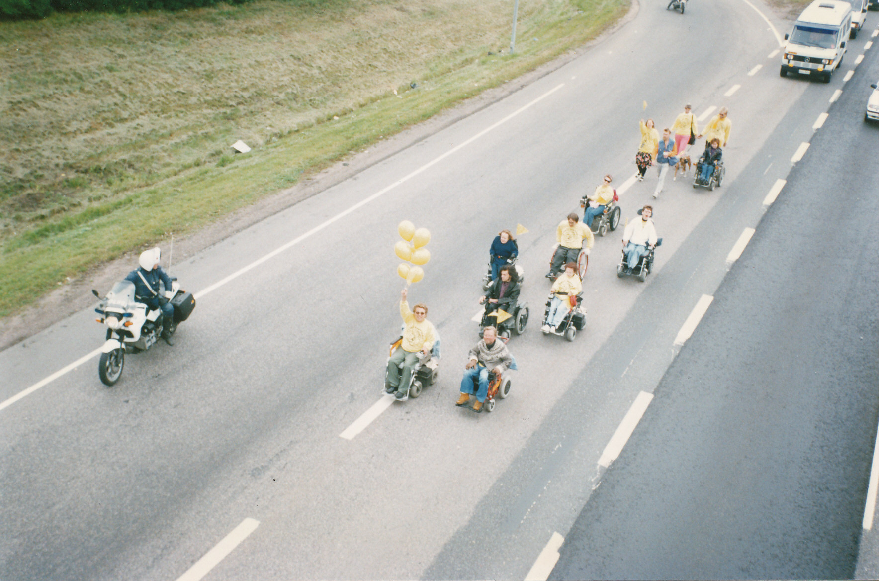 A color photograph of people rolling wheelchairs and walking on the highway, escorted by motorcycle police.